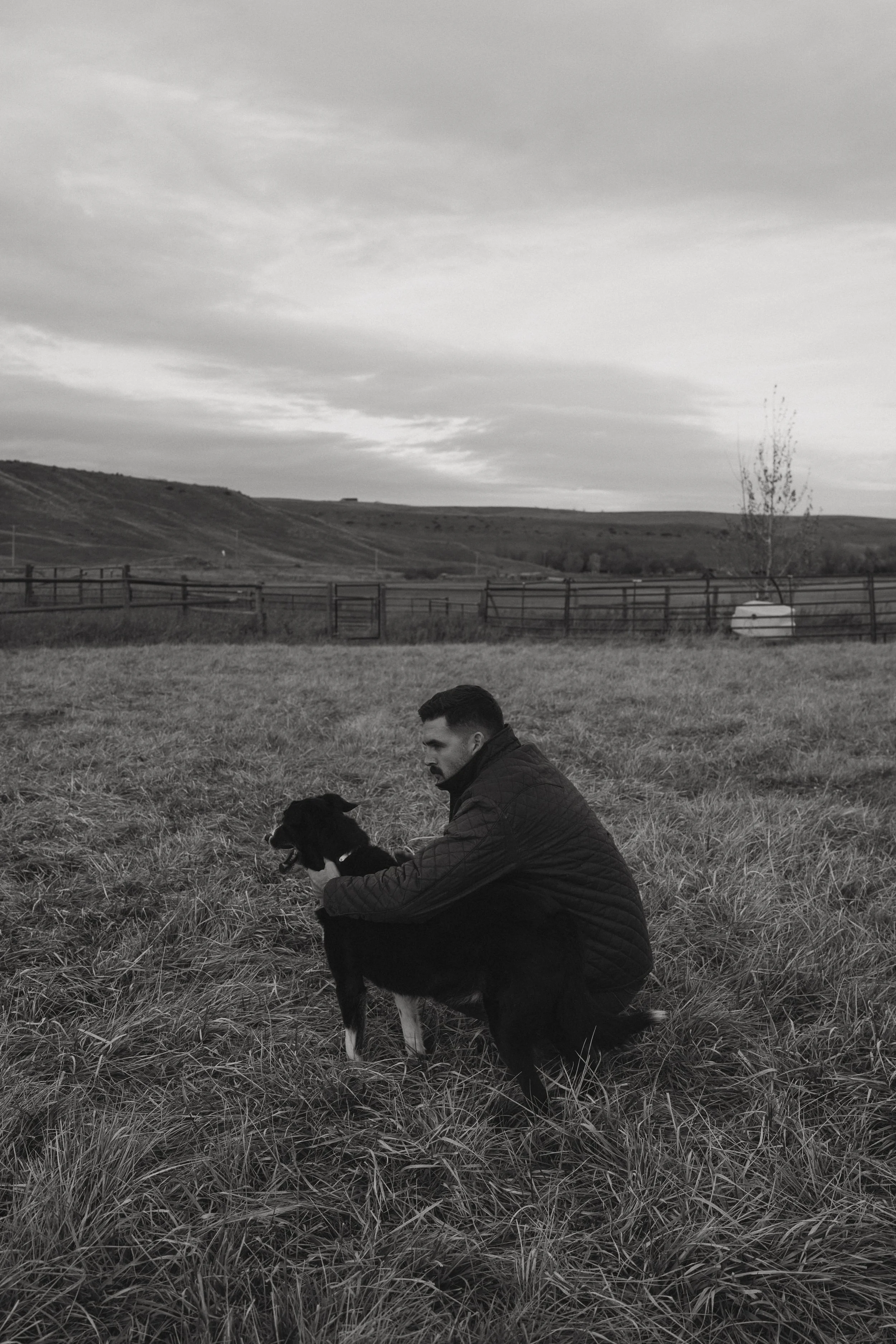 A man kneeling on the grass with a dog, in a rural landscape with rolling hills, a tree, and a wooden fence, under a cloudy sky.