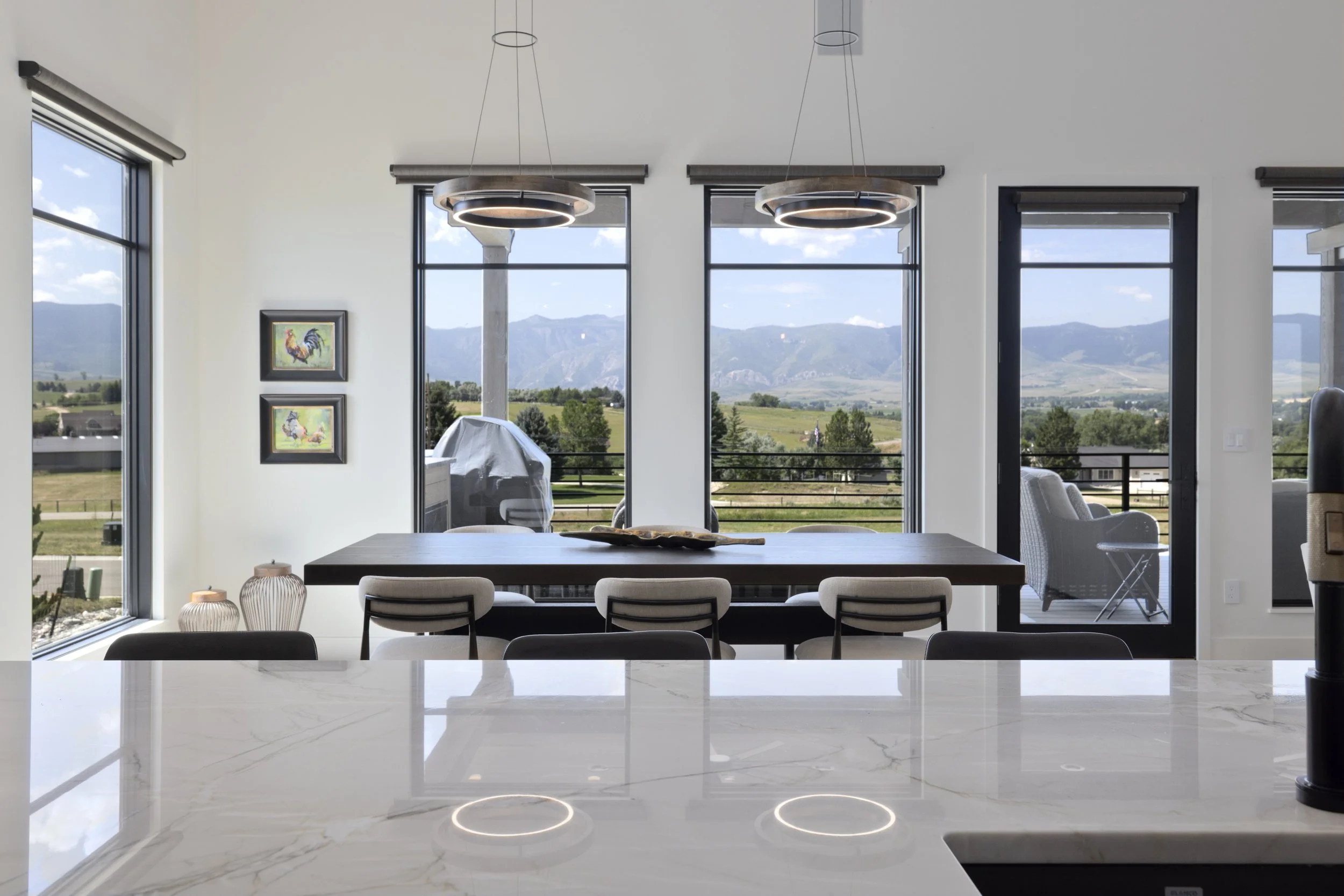 Modern kitchen with a white marble island countertop, dining table with chairs, large windows showing a scenic mountain view, and contemporary pendant lights.