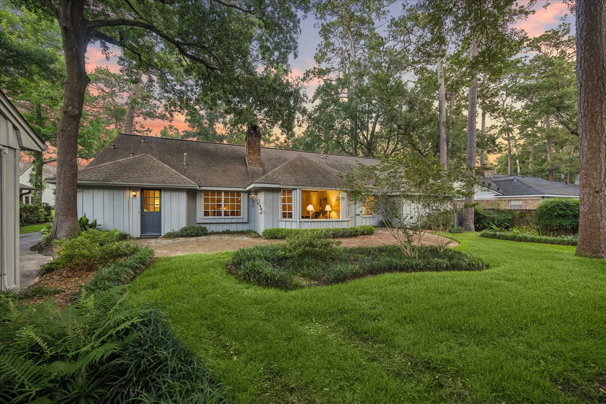 View of a house with a well-lit interior, surrounded by green lawn, foliage, and tall trees, during sunset.