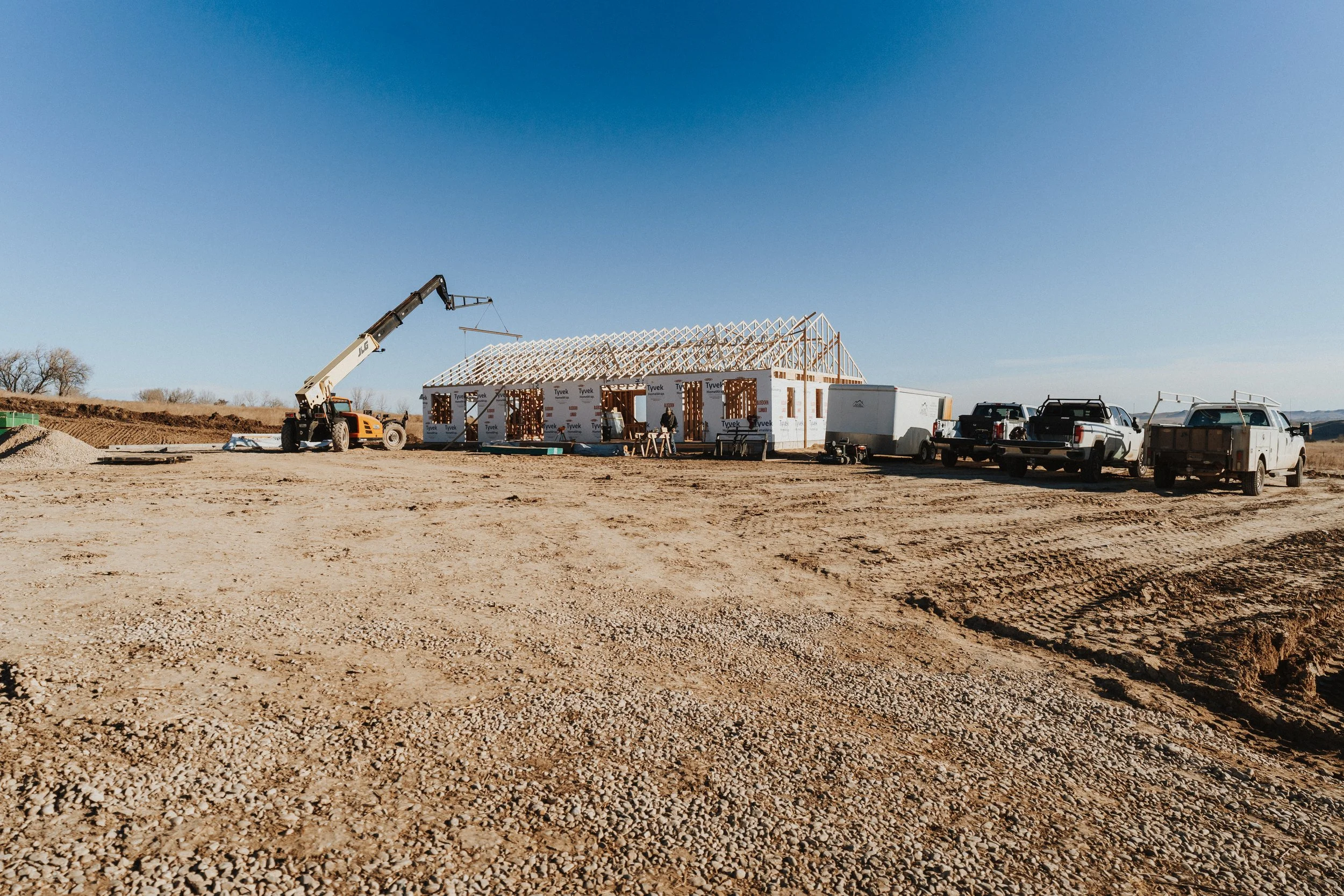 Construction site with a wooden house frame, construction vehicles, and workers under a clear blue sky.