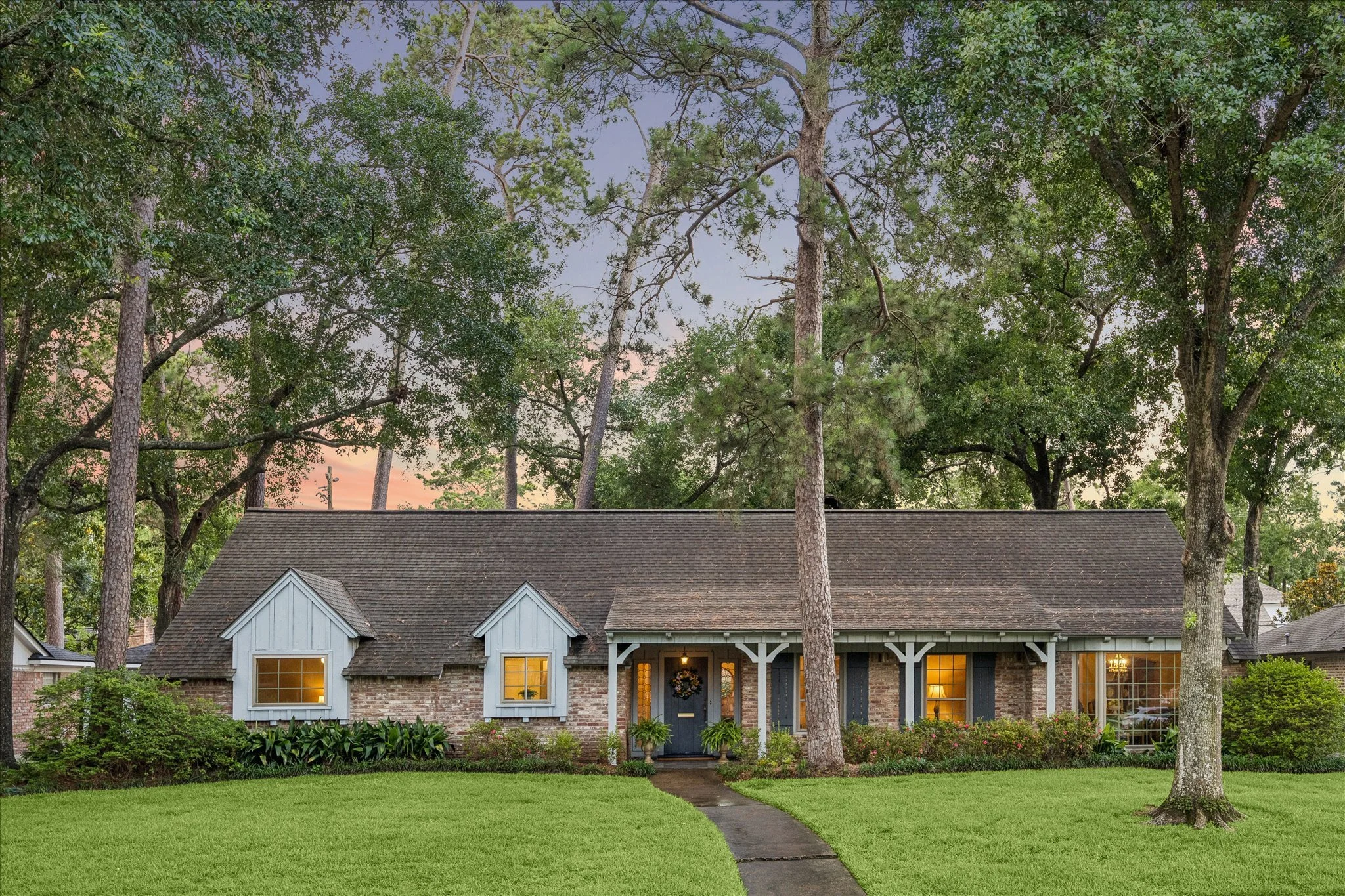 Front view of a cottage house with white trim, illuminated windows, and a pathway leading to the front door, surrounded by lush green grass and trees at sunset.