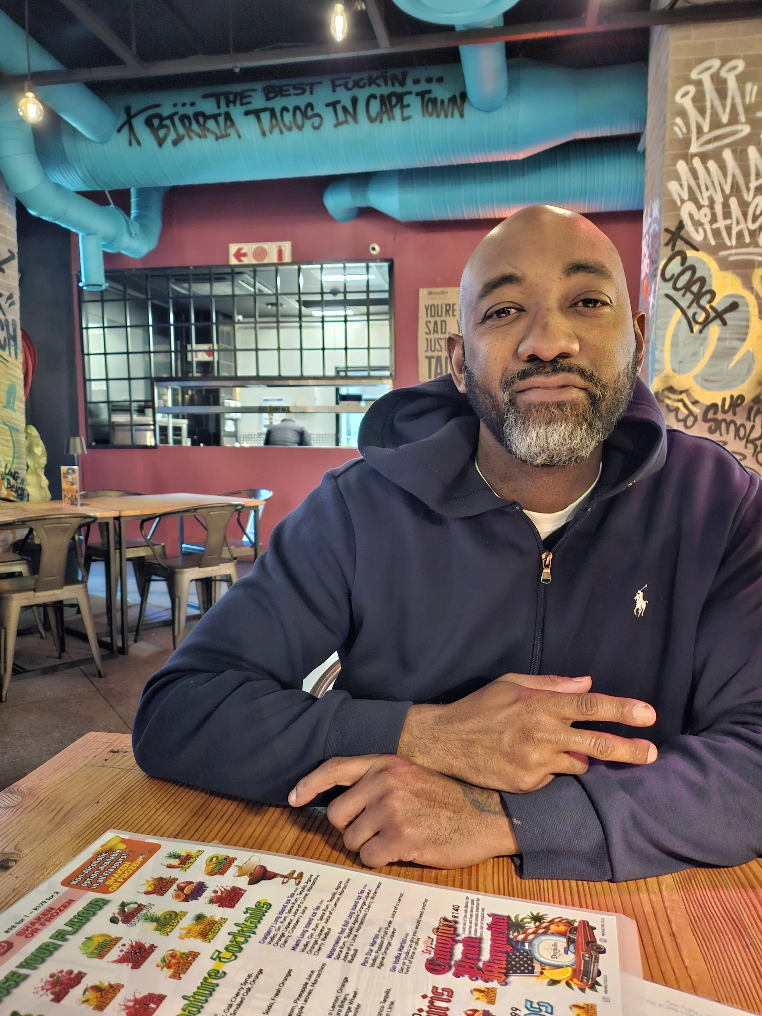 A man with a beard and mustache sitting at a wooden table in a restaurant, with a colorful menu in front of him. The restaurant has graffiti-style art on the walls, exposed ventilation pipes, and hanging light bulbs. There is a large window behind the man, and some chairs can be seen in the background.