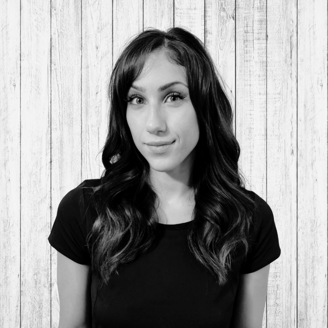Black-and-white portrait of a woman with wavy hair, slight smile, wearing a black top, against a wooden plank background.