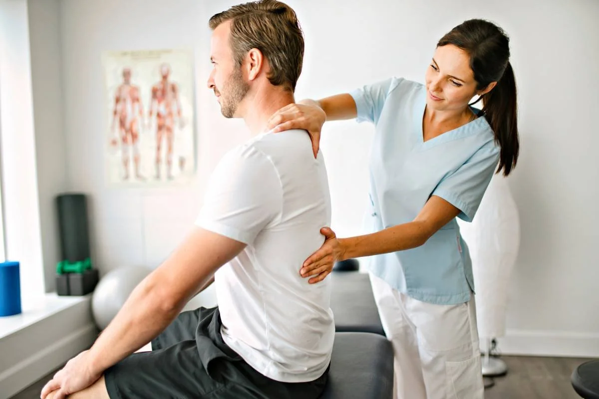 A healthcare professional, a woman in blue scrubs, providing chiropractic care to a man seated on an examination table, focusing on his shoulder.