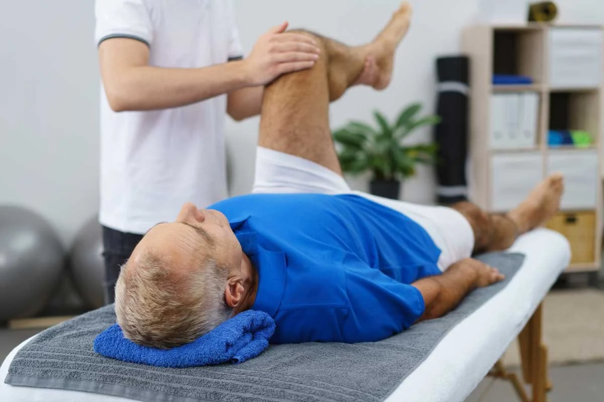 A physical therapist assisting an elderly man with leg exercises on an examination table in a clinic or therapy room.