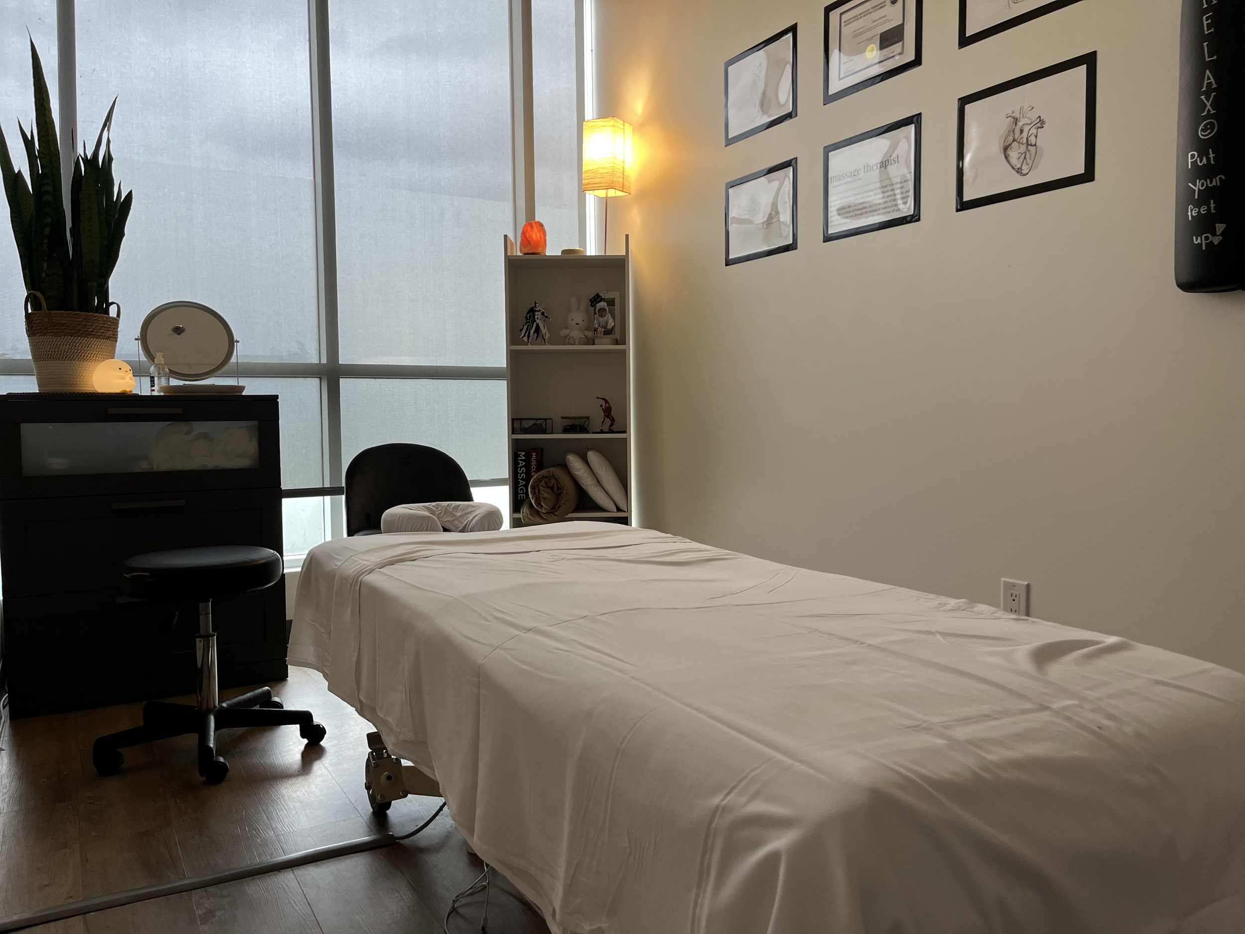 A massage therapy room with a massage bed, black chair, a small black cabinet with plants and decor, a white bookshelf with toys and books, framed certificates, and a large window with pull-down shades.