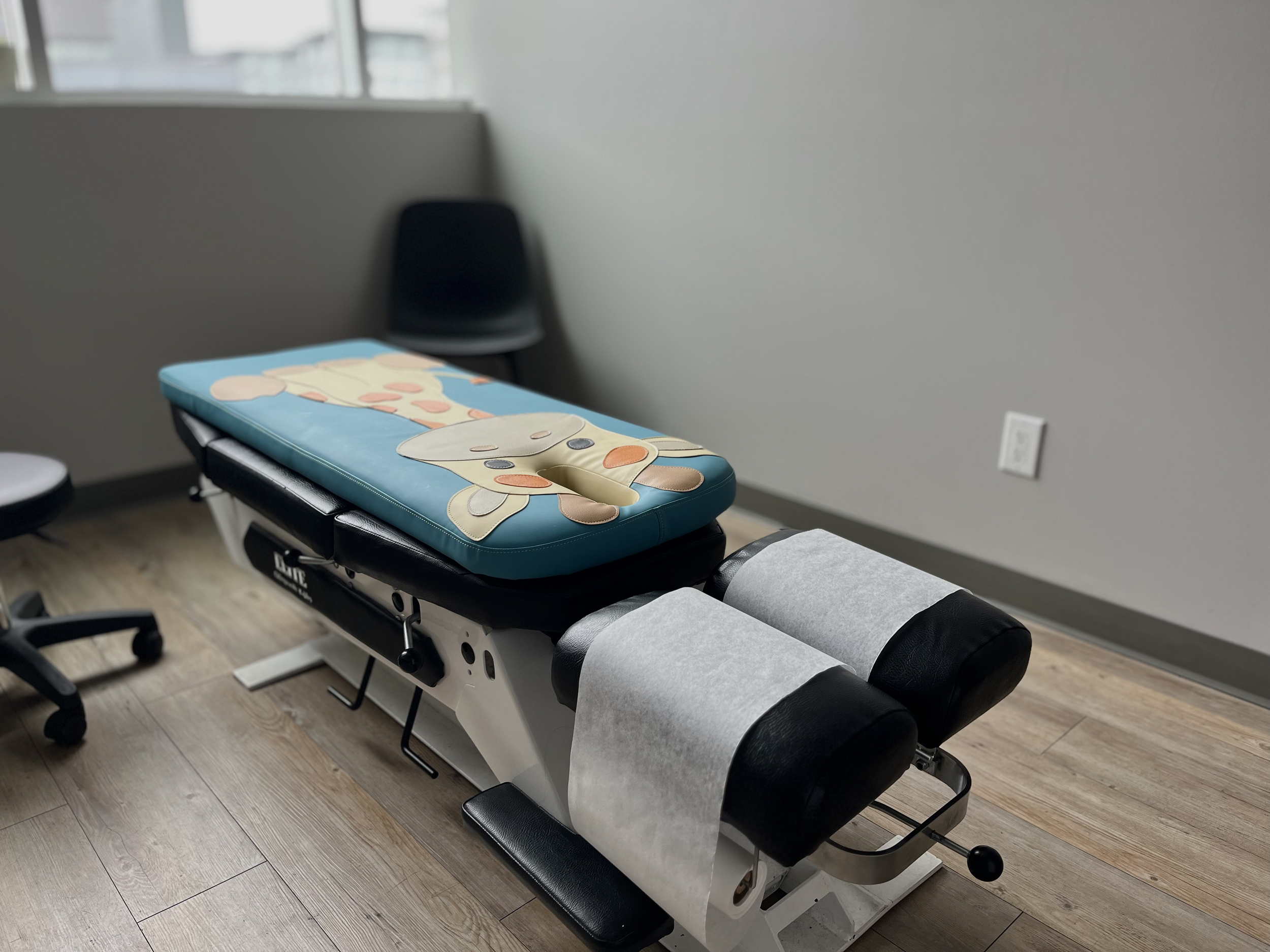 Medical or therapy examination table with a cartoon cow pillow in a clinical room with grey walls and wooden floor.