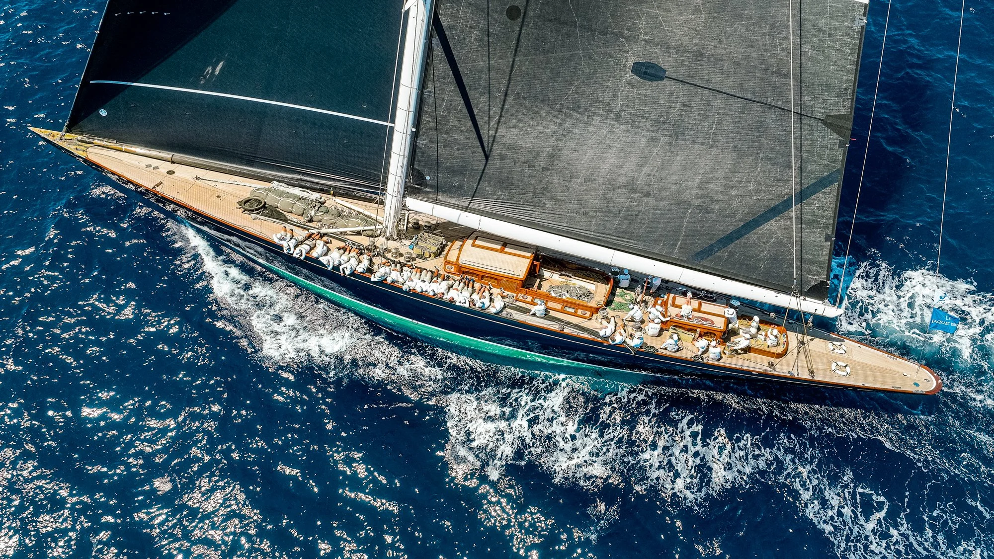 Vue aérienne d'un voilier de course naviguant en mer avec des équipages à bord, vue du dessus du bateau et de la mer bleue en contrebas.