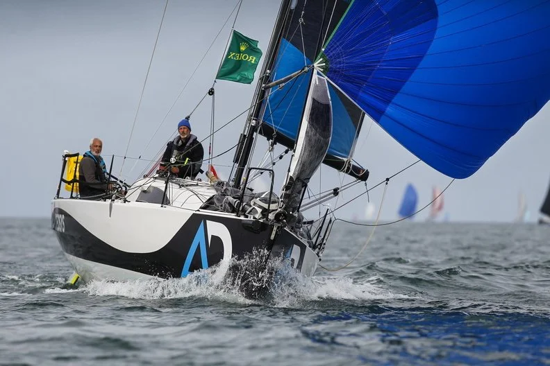 Deux hommes naviguent un voilier de course avec des voiles bleues, une enrouleur et un drapeau vert avec l'inscription 'ROLEX'.