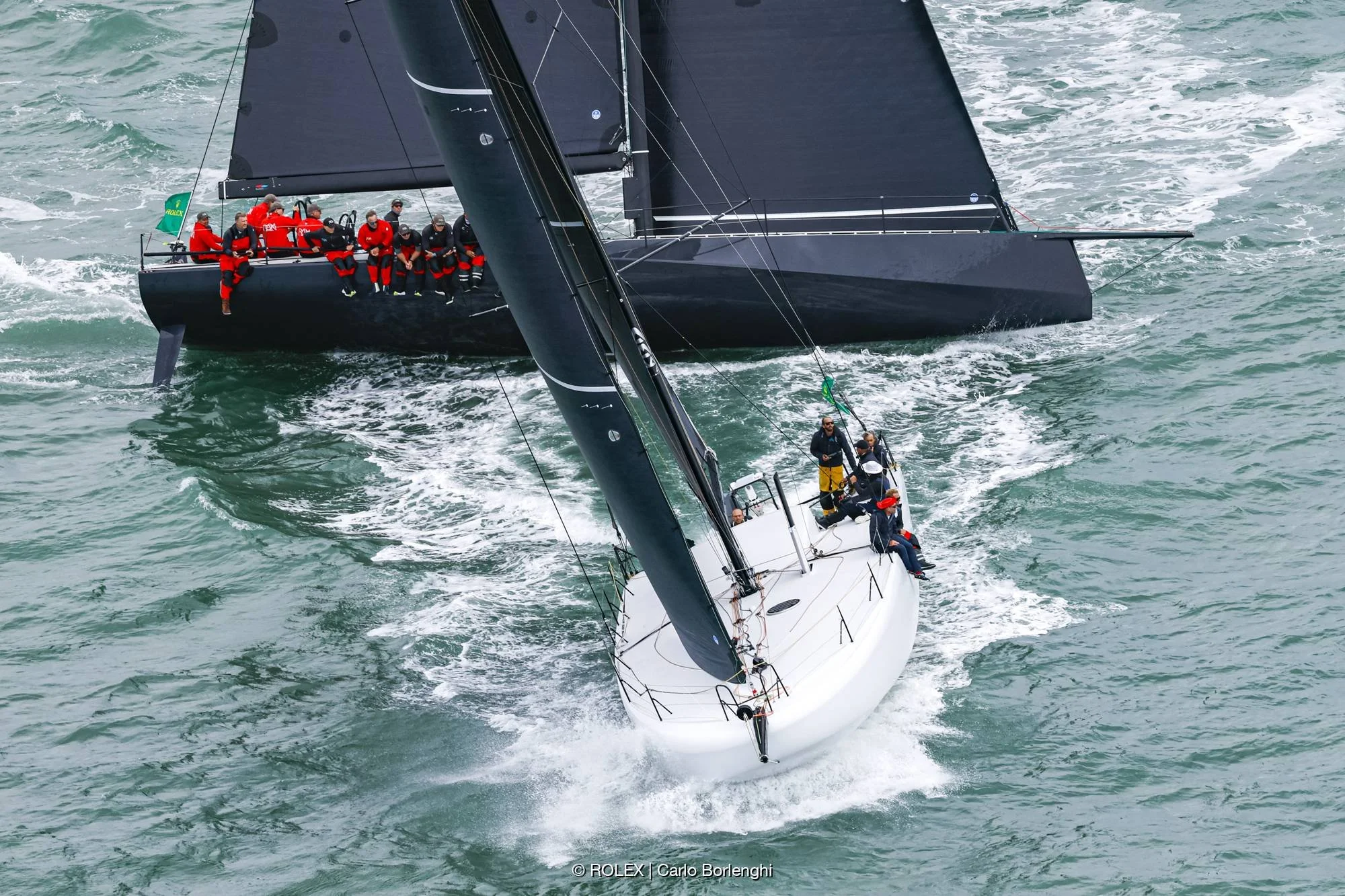 Deux yachts de course naviguent rapidement sur la mer, avec plusieurs personnes à bord, portant des vêtements de sport et de sécurité. Les yachts ont des coques blanches et noires avec des voiles noires.