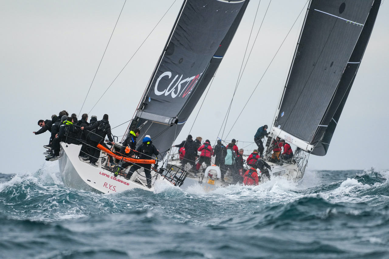 Voilier de course avec plusieurs équipiers naviguant dans des eaux agitées par vent fort, sous un ciel nuageux.