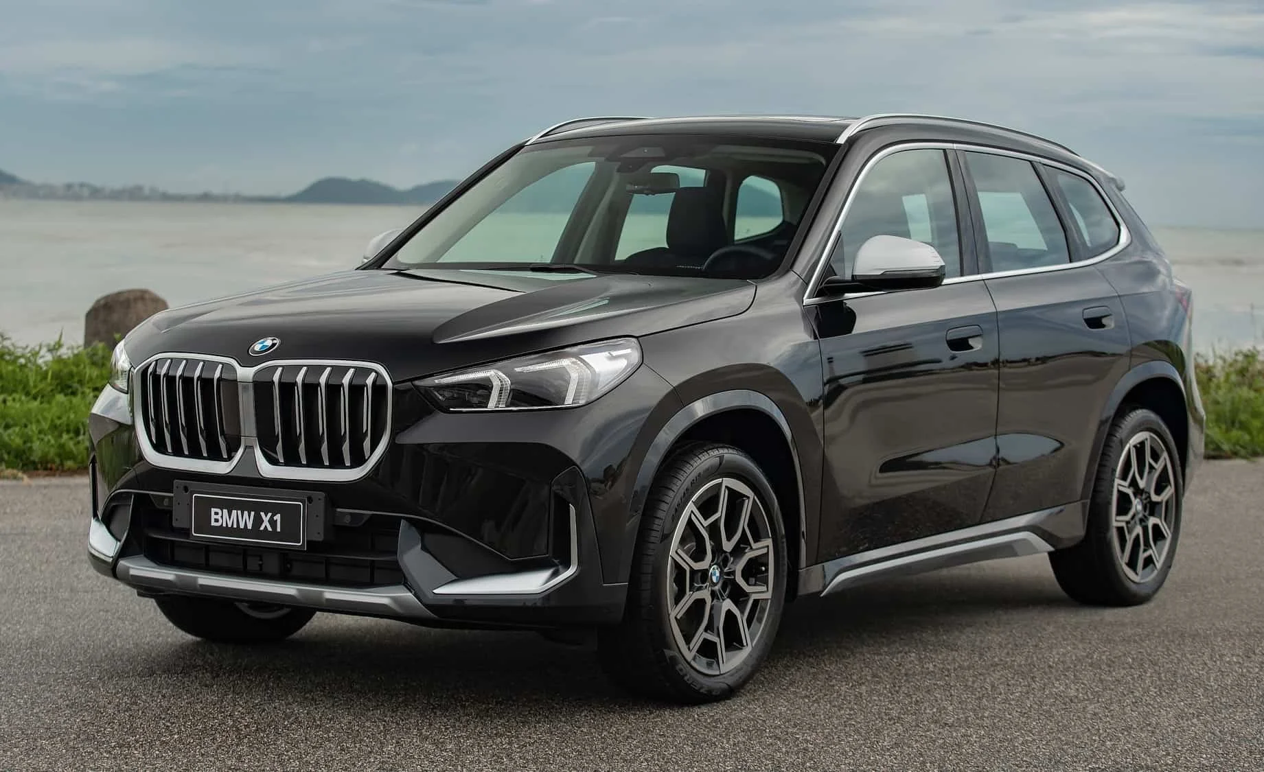 Black BMW X1 SUV parked on an asphalt road near the coast with water and hills in the background.