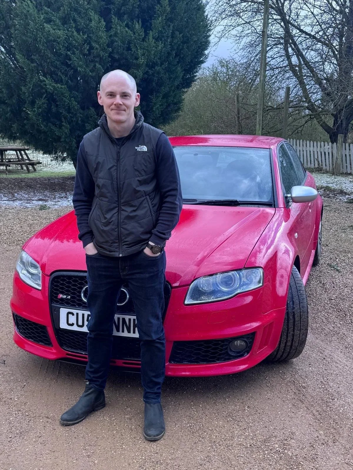 A man standing in front of an Audi B7 RS4 car on a gravel driveway, with trees and a fence in the background.