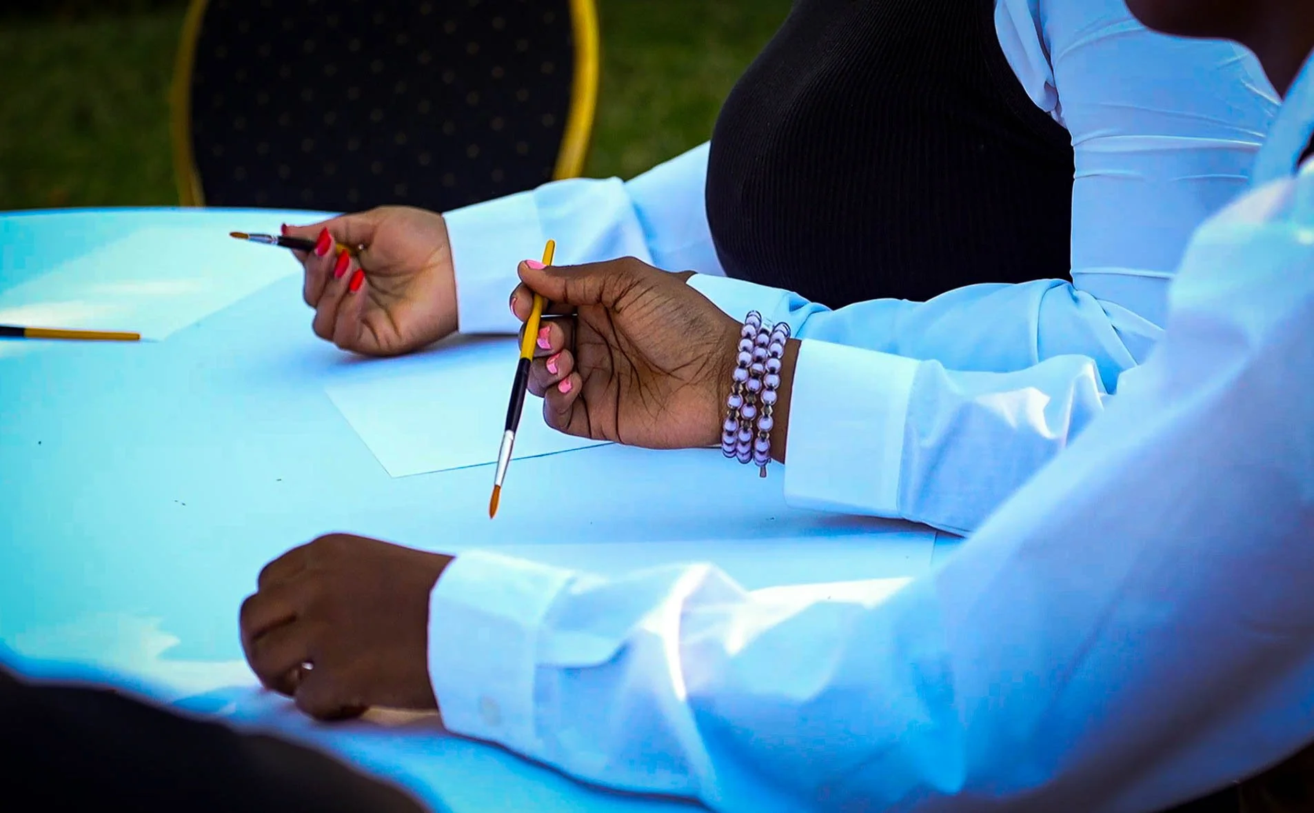 Close-up of two people sitting at a white table, holding paintbrushes, with one person wearing beaded bracelets and the other with bright pink painted nails.