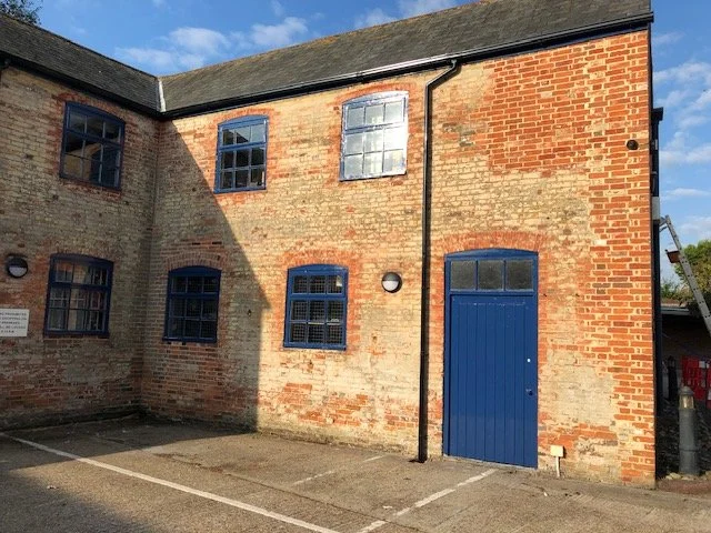 A brick building with five windows painted blue and a blue door, situated in a parking lot with marked parking spaces, under a partly cloudy sky.