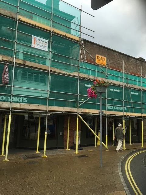 Construction scaffolding covered with green netting outside a McDonald's restaurant, with a pedestrian walking on the sidewalk and a flower basket hanging from a streetlight.