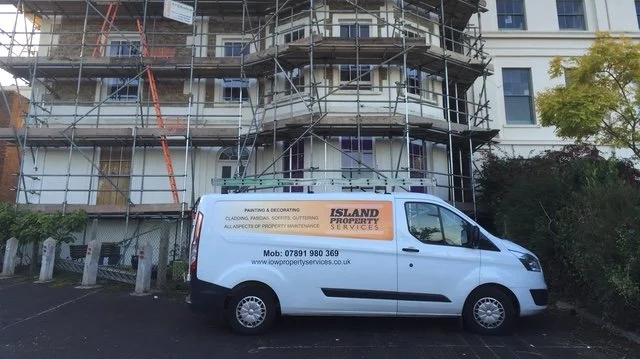 A white van with the signage 'Island Property Services' parked in front of a multi-story building under construction, with scaffolding surrounding the building.