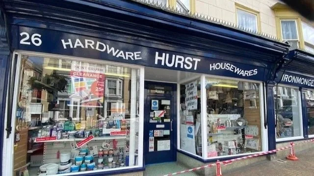 Outside view of a hardware store named 'Hurst Housewares' with a blue awning and large display windows showcasing tools and supplies.