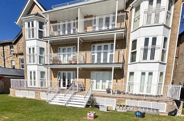Three-story brick apartment building with white balconies and railings, front steps leading to entrances, and a grassy yard with scattered objects.