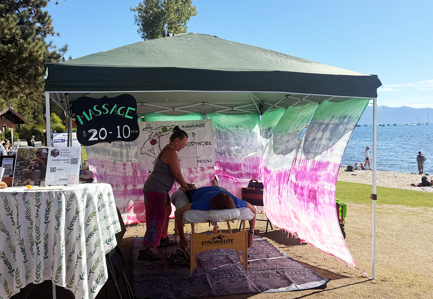A woman receives a massage outdoors under a canopy by a lake with boats and mountains in the background.