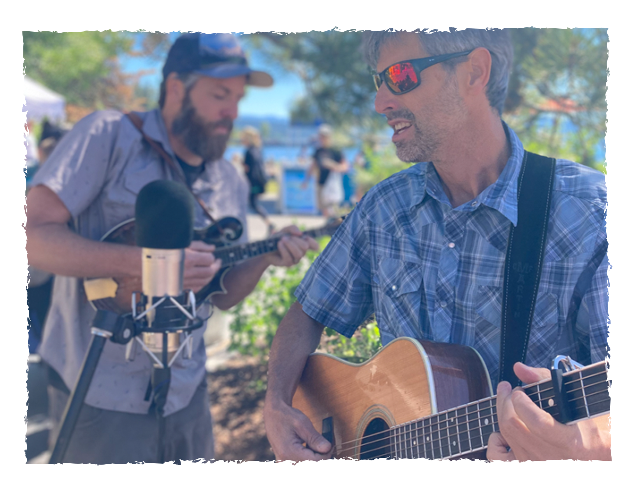 Two men playing guitars and singing outdoors, with one man in focus wearing sunglasses and the other blurred playing a mandolin, surrounded by trees and people.