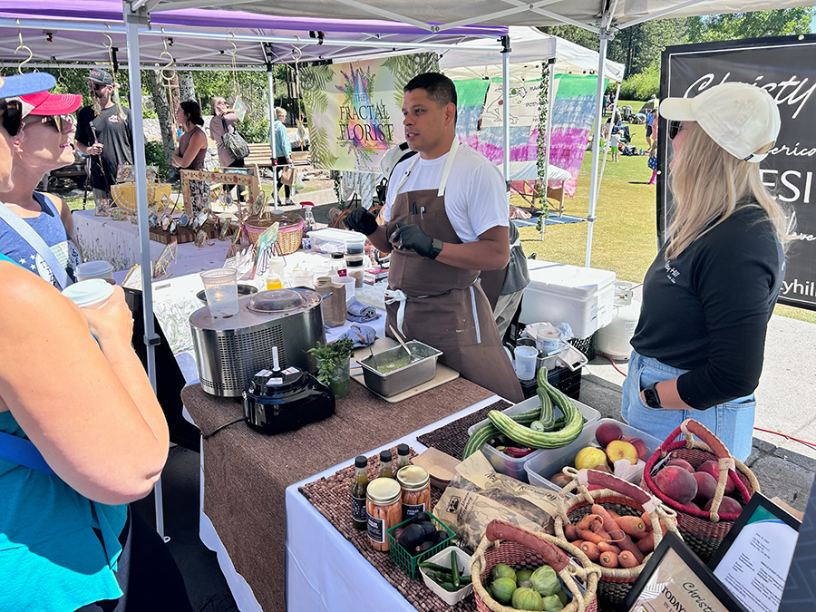 Vendor at a market booth demonstrating cooking with fresh vegetables, including zucchini and carrots, with a crowd of shoppers nearby.