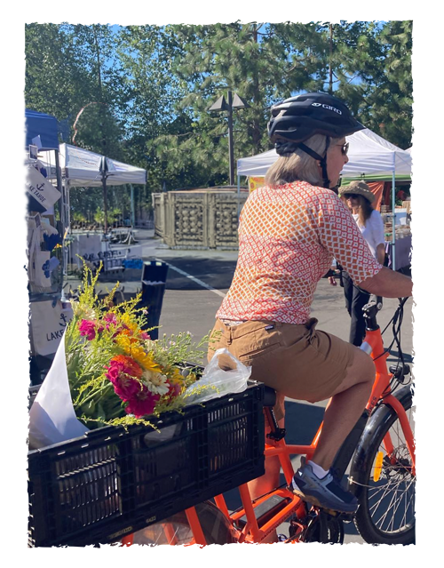 Woman wearing a helmet, orange patterned shirt, and brown shorts riding an orange electric bike with a basket full of colorful flowers at an outdoor market or event on a sunny day.