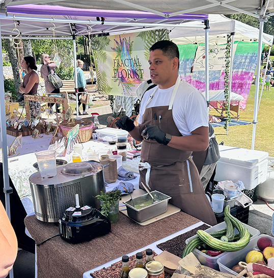 Vendor at a market booth demonstrating cooking with fresh vegetables, including zucchini and carrots, with a crowd of shoppers nearby.