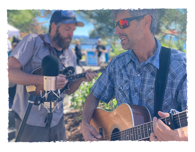 Two men playing acoustic guitars outdoors, with one wearing sunglasses and the other wearing a cap, during daytime.
