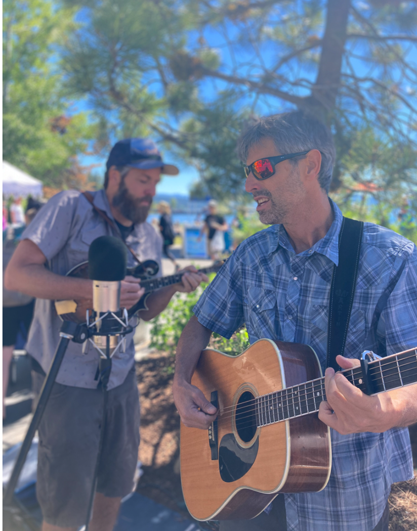 Two men playing guitars and singing outdoors, with one man in focus wearing sunglasses and the other blurred playing a mandolin, surrounded by trees and people.