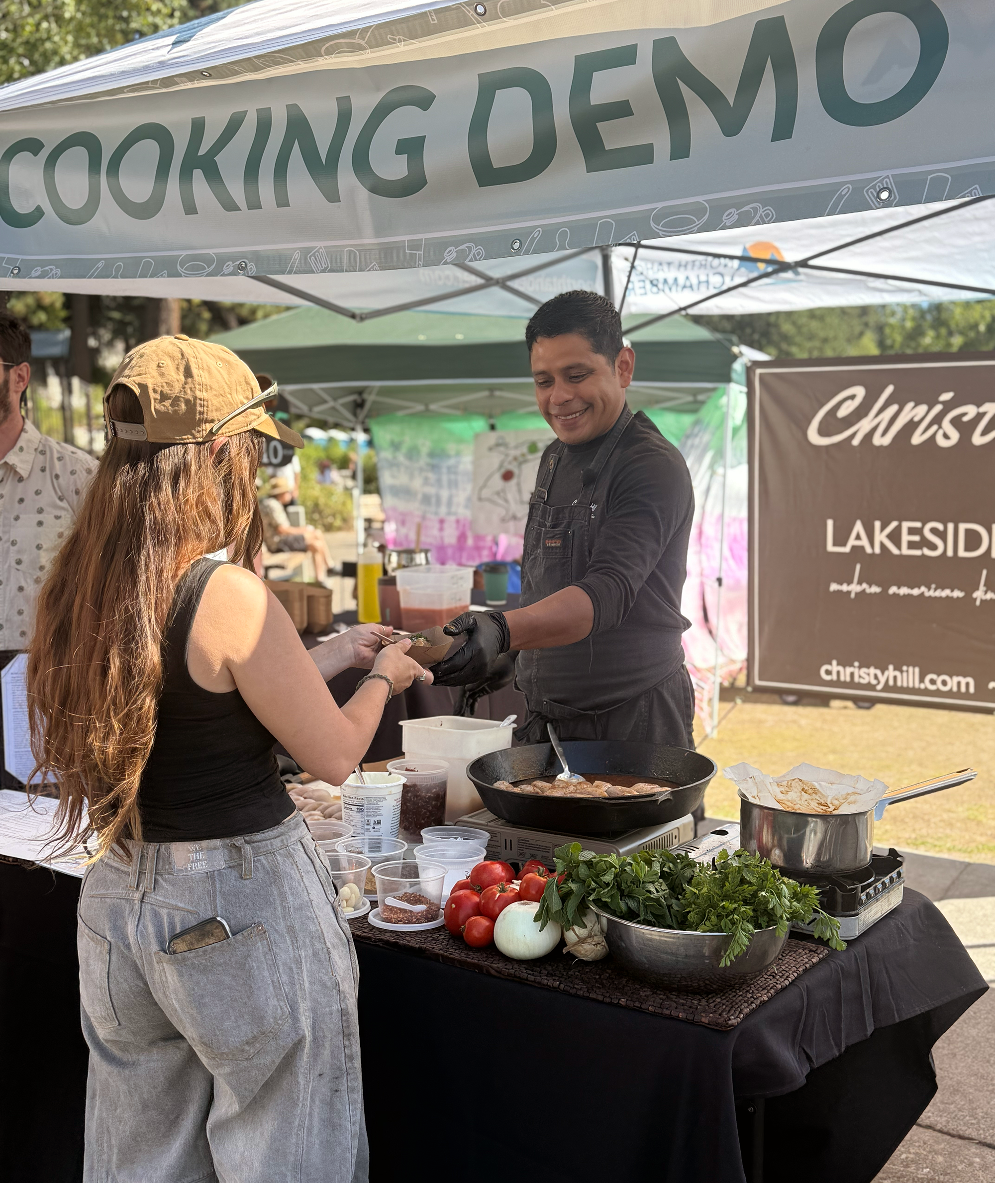 Vendor at a market booth demonstrating cooking with fresh vegetables, including zucchini and carrots, with a crowd of shoppers nearby.