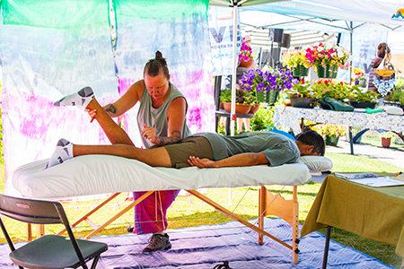 A woman receives a massage outdoors under a canopy by a lake with boats and mountains in the background.
