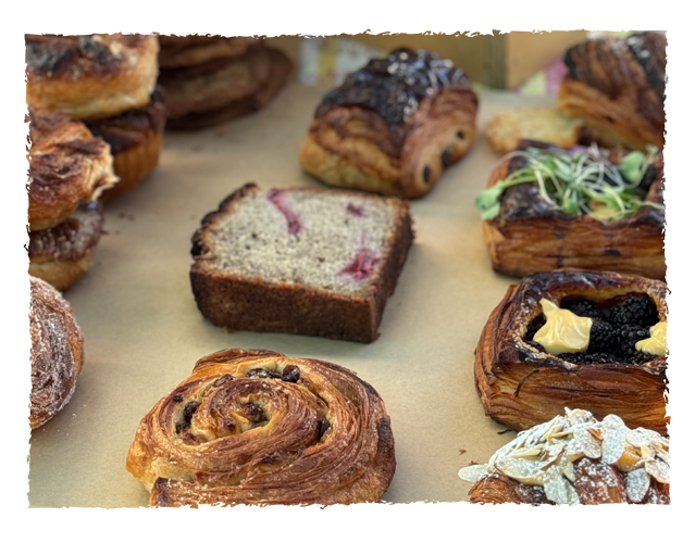 Assorted baked goods including a slice of marble cake, cinnamon roll, chocolate croissant, fruit Danish, and other pastries on a baking sheet.