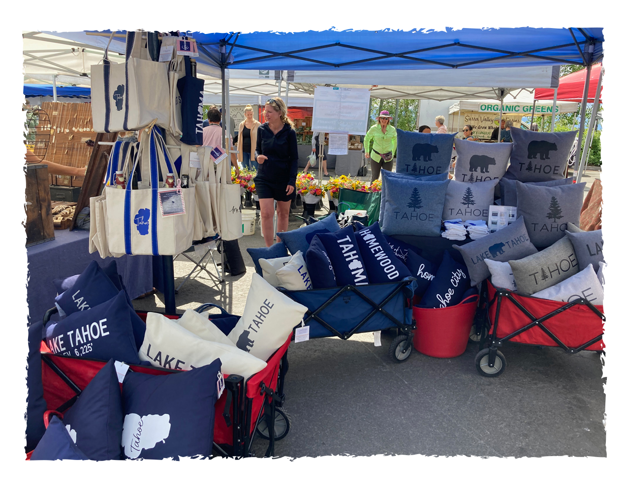 Outdoor market booth selling pillows and tote bags with Tahoe and Lake Tahoe designs, with shoppers browsing in the background.
