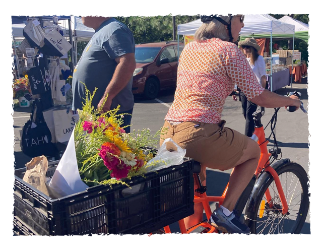 A woman riding an orange bike with a basket filled with bright flowers at an outdoor market. Other market stalls and people are visible in the background.