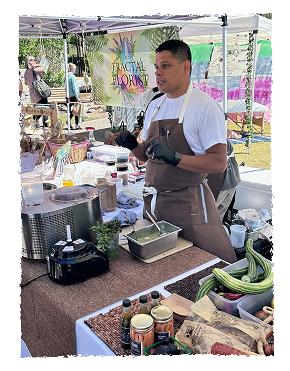 A man wearing a white t-shirt, apron, and black gloves preparing food at a booth called The Fractal Florist, which has a colorful banner and various jars and fresh produce on the table outside at a market.