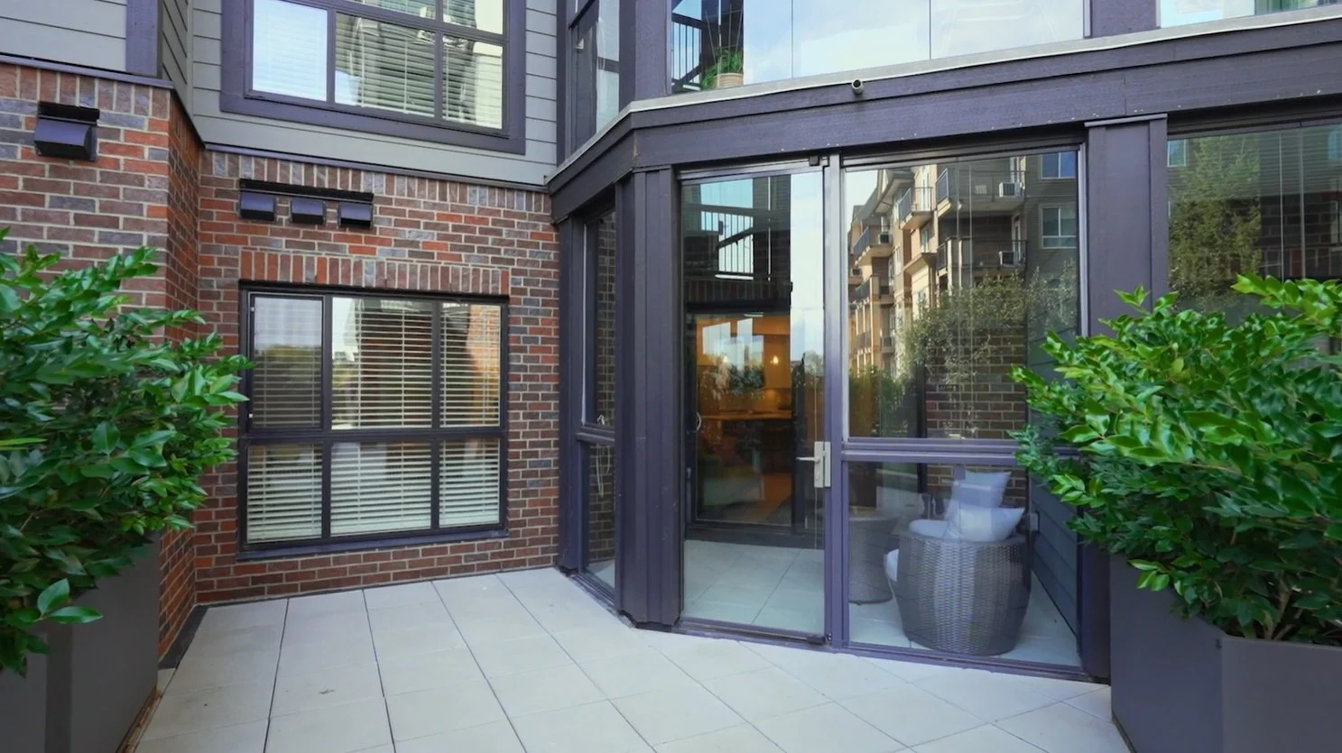 Outdoor patio area with two large plants in gray planters, brick and glass building walls, and a sliding glass door leading inside.