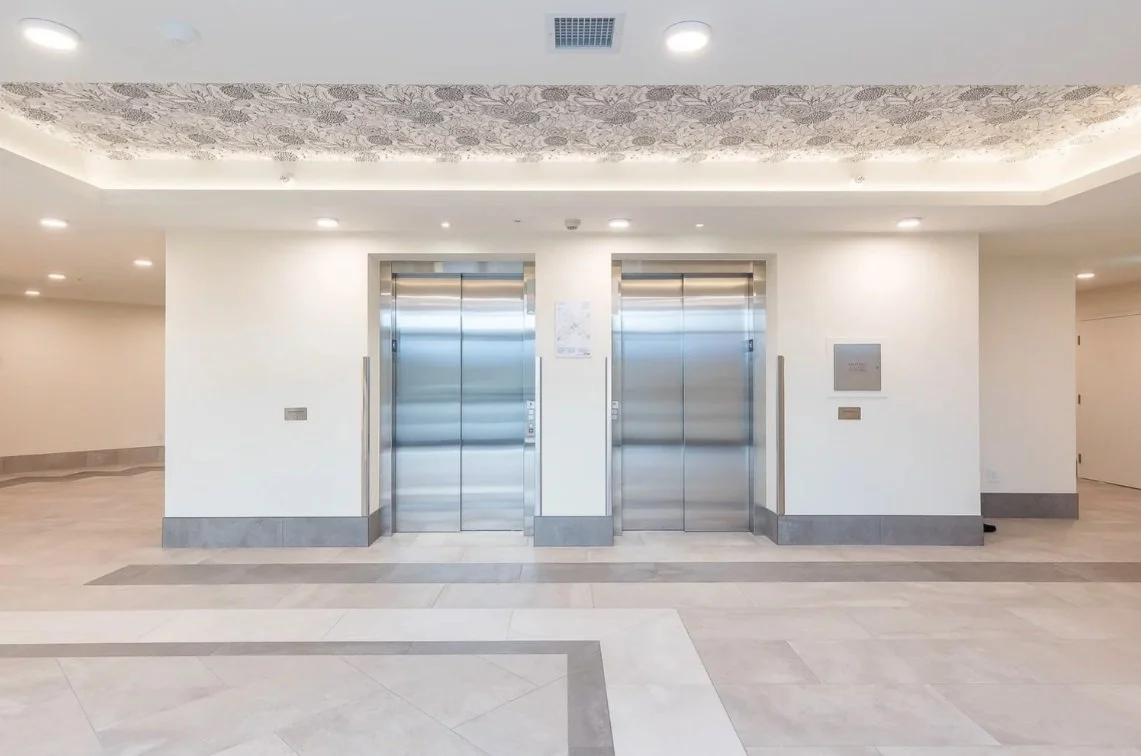 Modern indoor space with two stainless steel elevator doors, white walls, and ceiling lighting, including decorative ceiling detail.