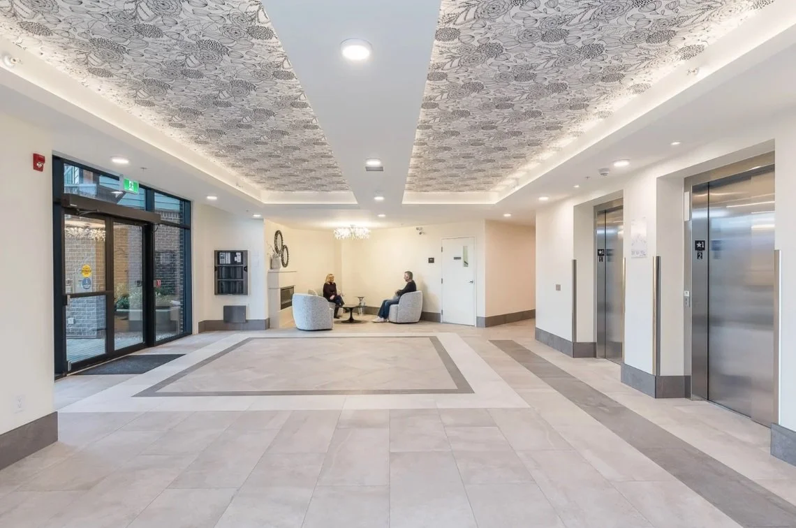 Lobby area with two people seated in conversation, elevator doors on the right, glass entrance doors on the left, decorative ceiling, and neutral-colored flooring.
