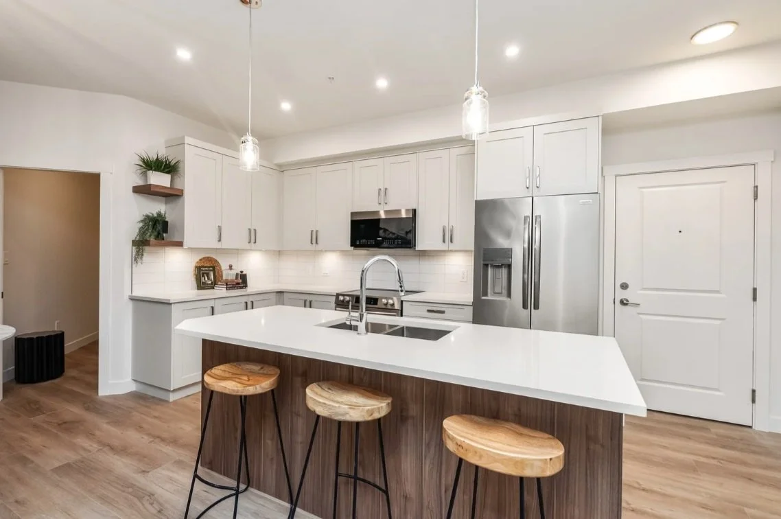 Modern kitchen with white cabinets, stainless steel appliances, a white island with a wooden base, and three wooden barstools. Recessed lighting and pendant lights above the island.
