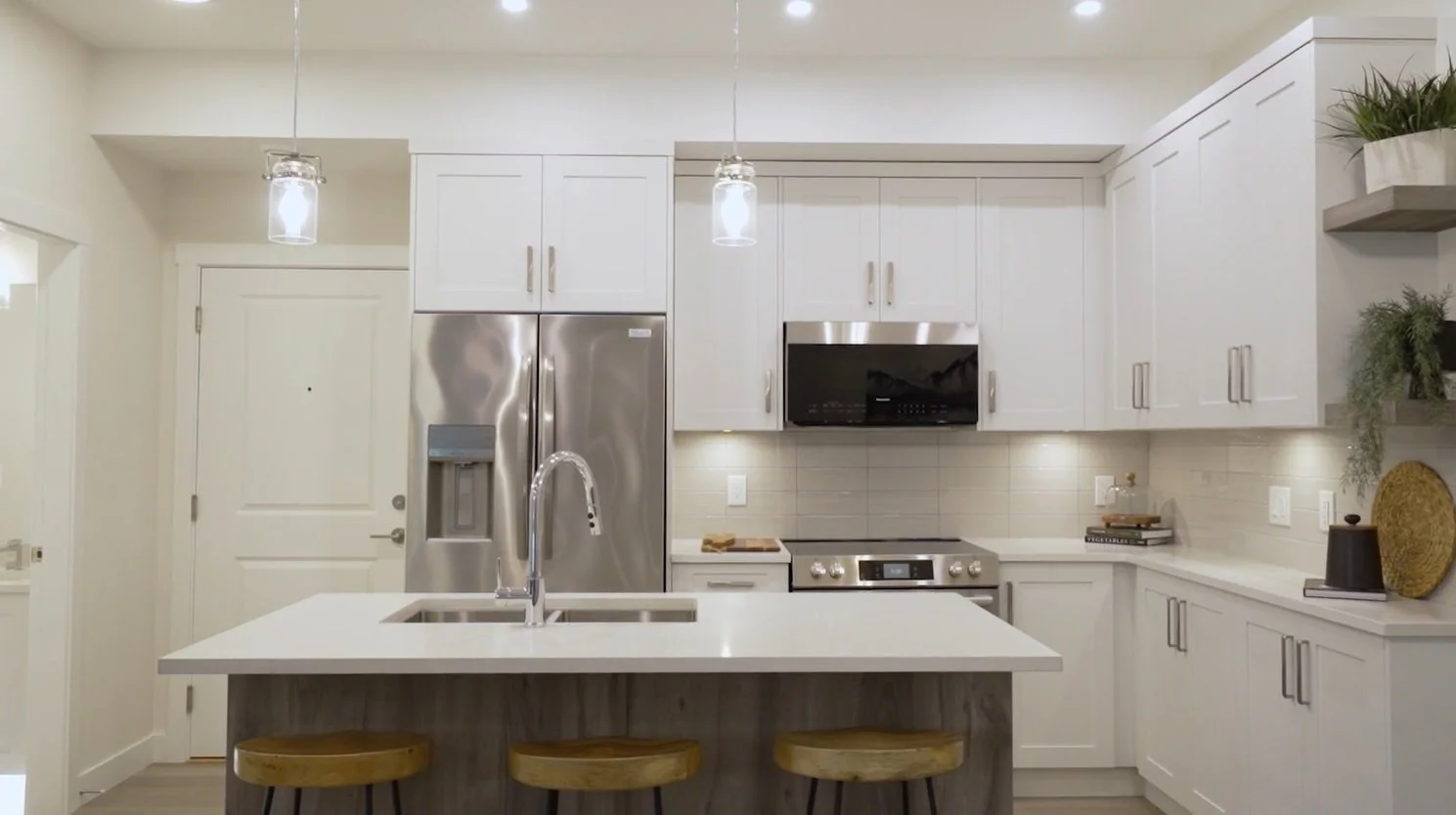 Modern white kitchen with stainless steel refrigerator, microwave, and oven, island with sink and wooden stools, pendant lights, and decorative plants.