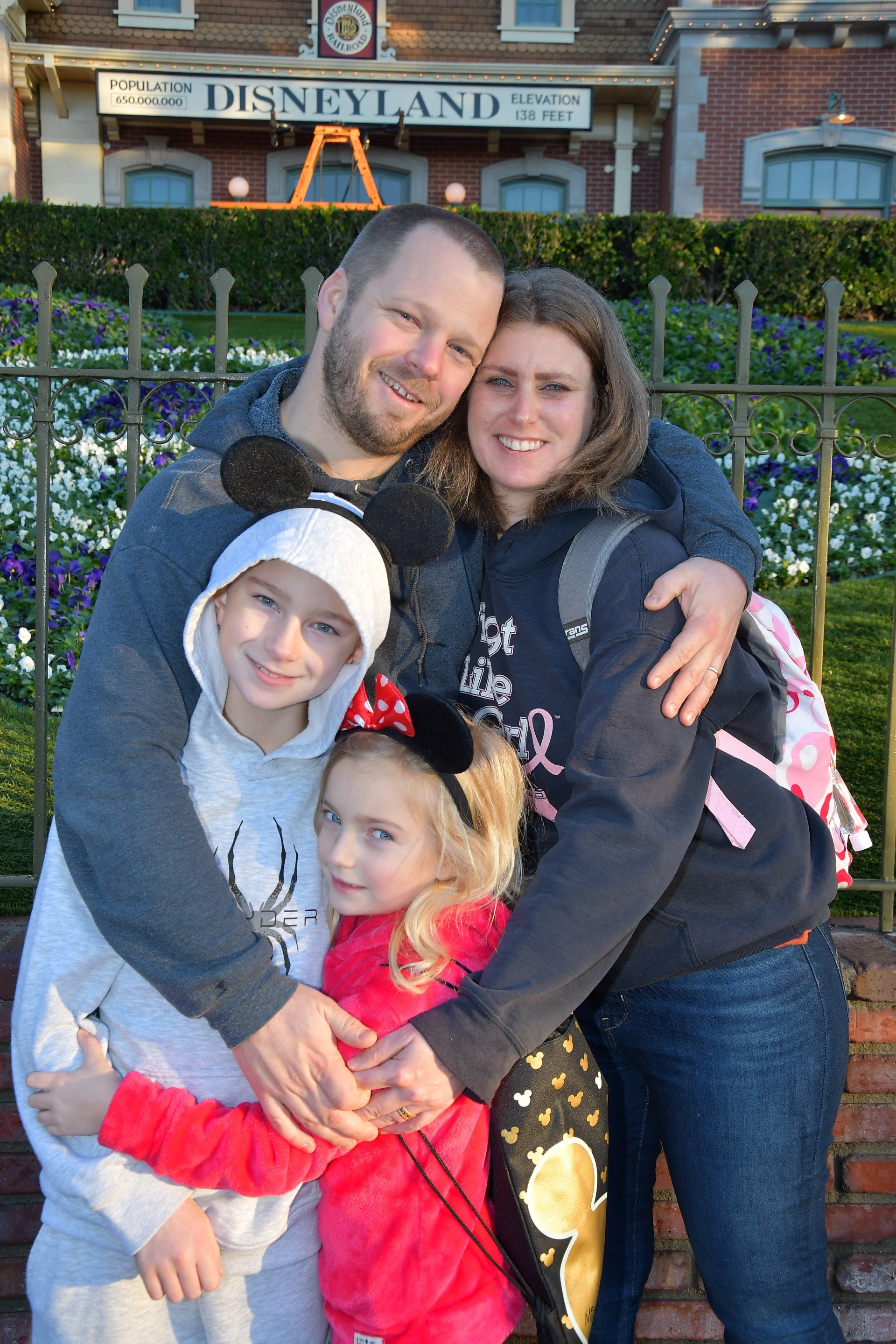 A family of four, wearing Disney-themed accessories, hugging in front of Disneyland with Flower Garden and Disneyland sign in the background.