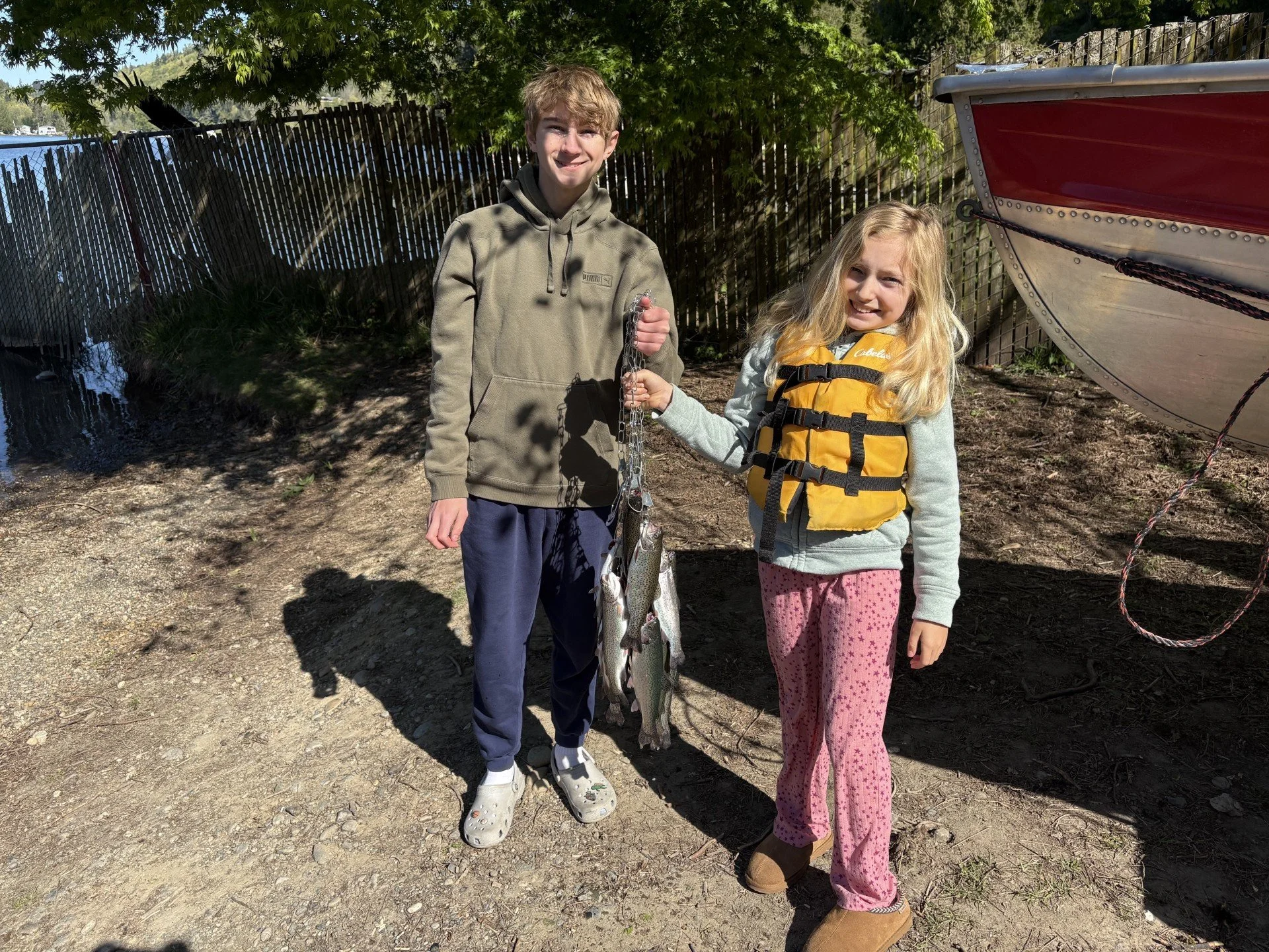 A boy and a girl standing outdoors next to a boat, holding a string of fish they caught, with a fence and trees in the background.