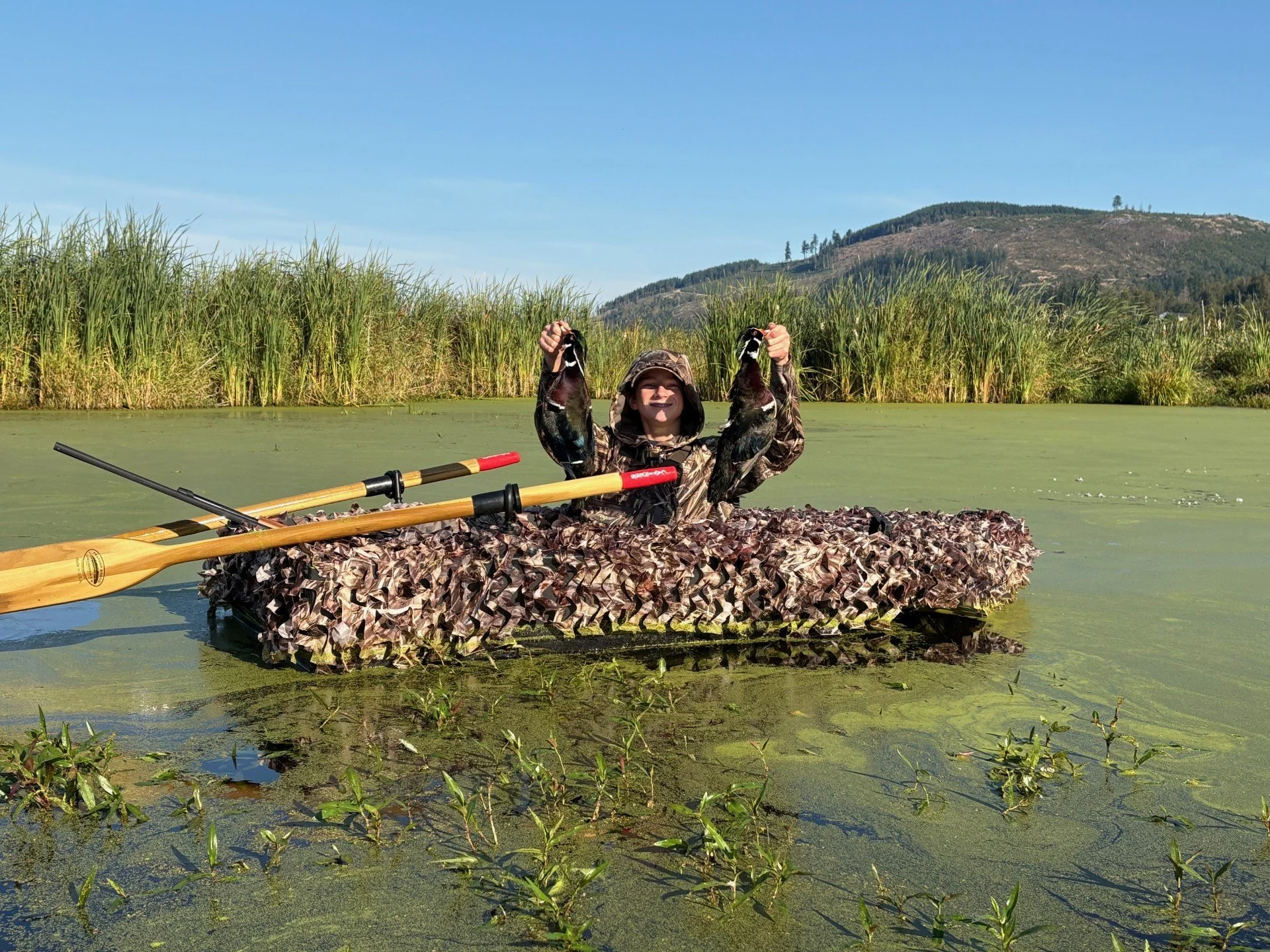 A young boy in camouflage clothing, sitting in a small boat covered with camouflage material, is holding two ducks aloft in a wetland with tall grasses and a mountain in the background.