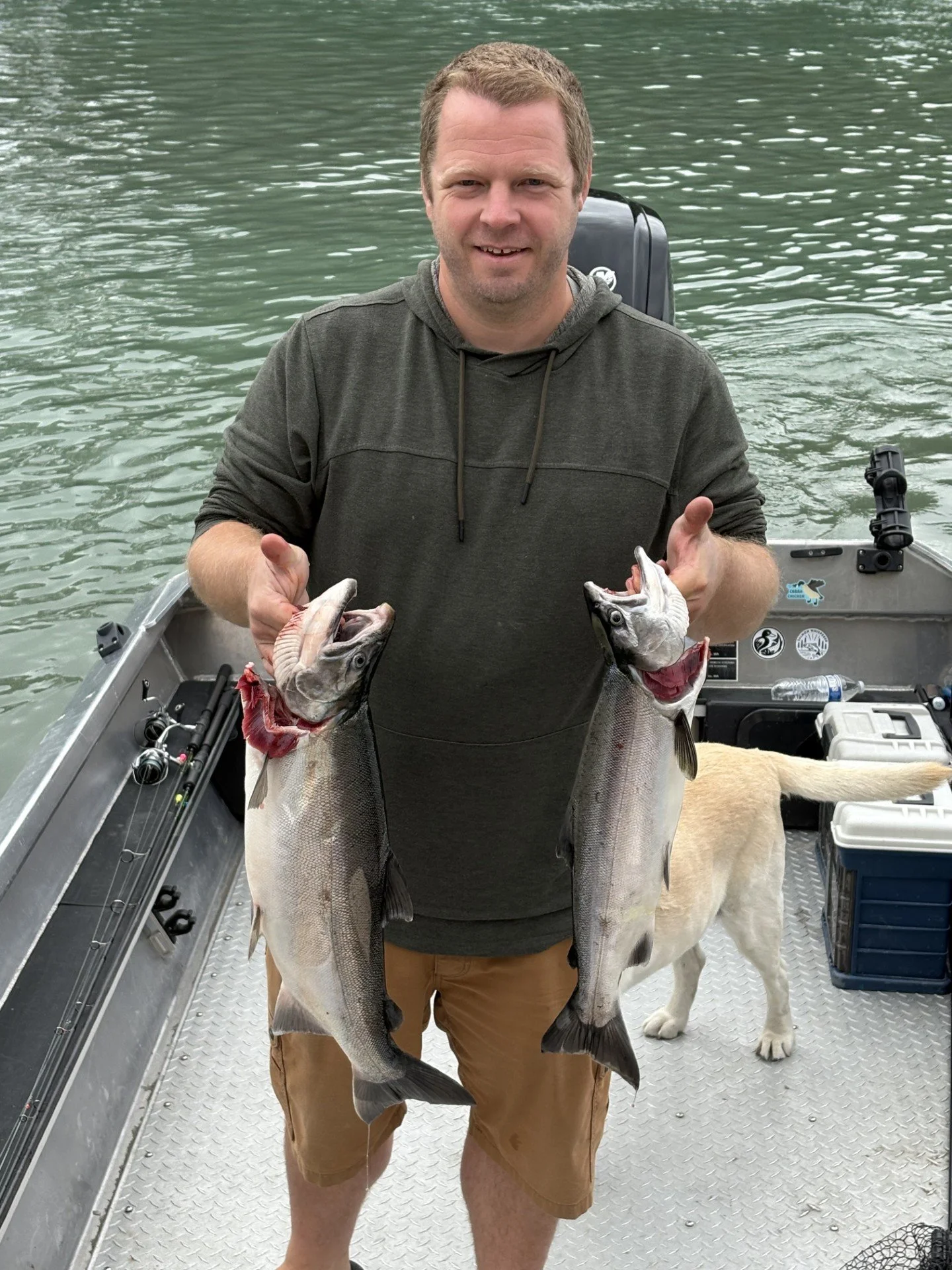 A man standing on a boat holding two large fish, with a dog in the background and fishing gear around.
