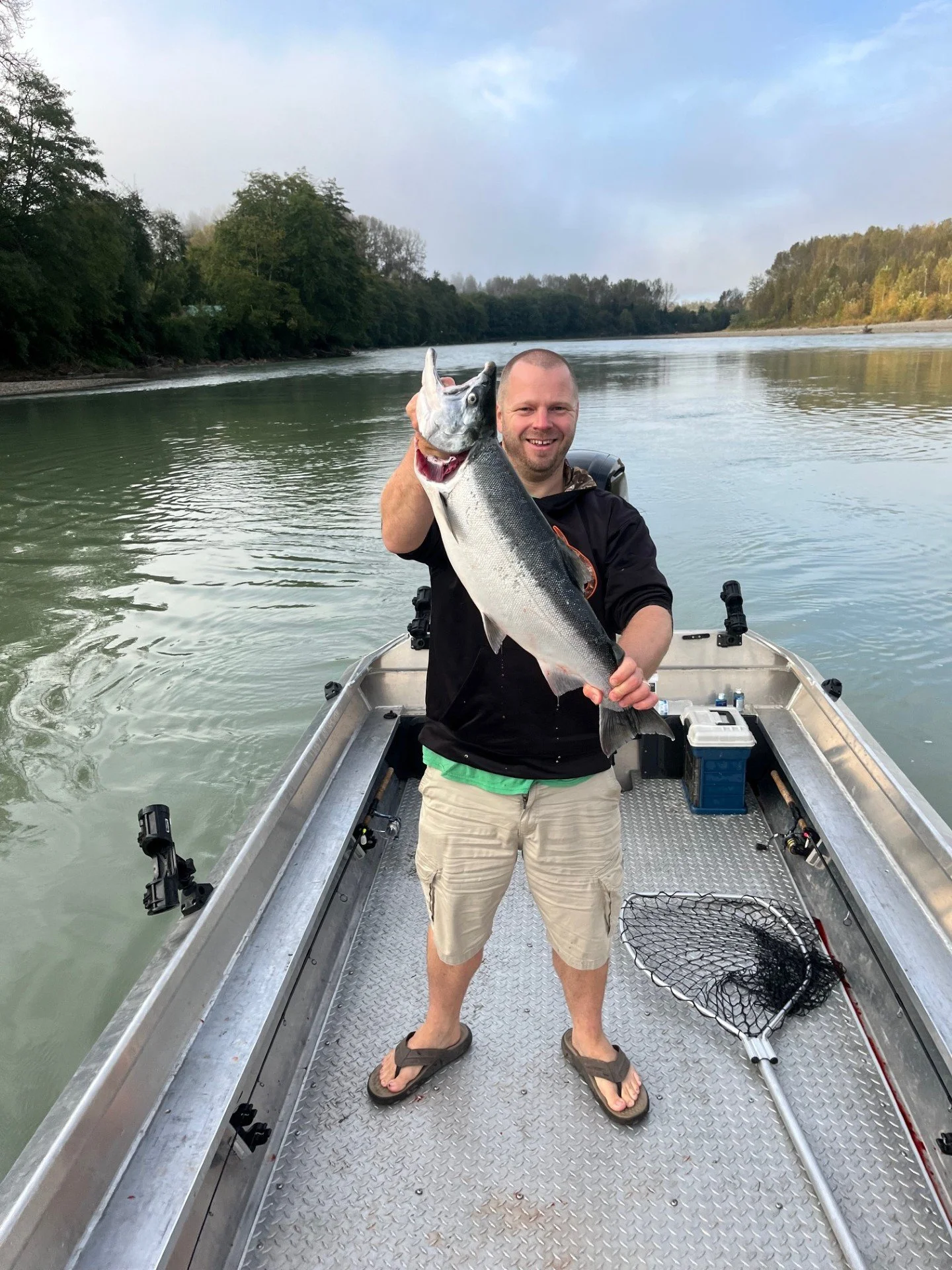 A man in a black hoodie and beige shorts standing on a boat, holding a large fish he caught, with water and trees in the background.