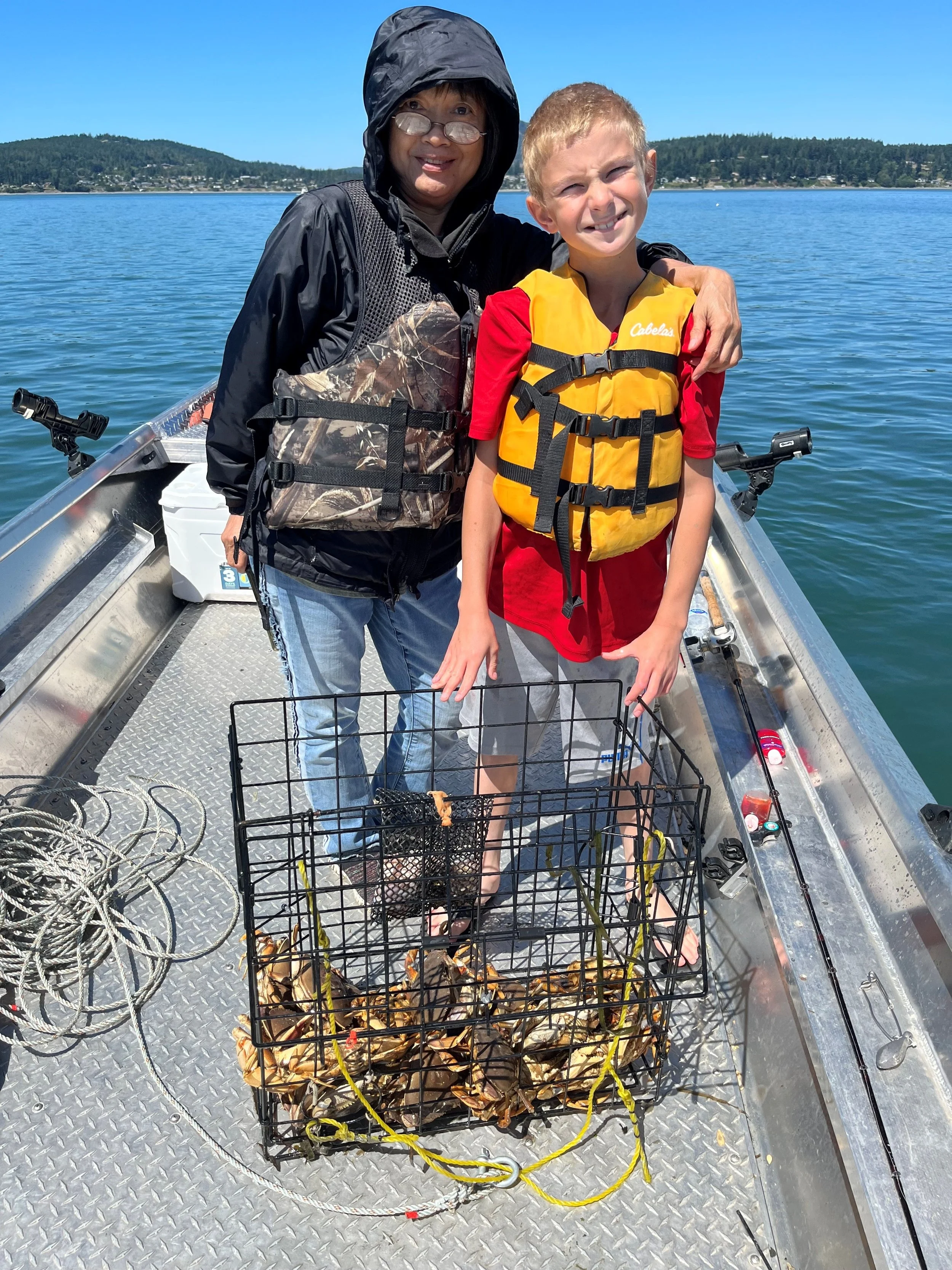 Two people, an adult and a child, on a boat with a basket of freshly caught crawfish, surrounded by ropes and fishing gear, with a lake and hills in the background.