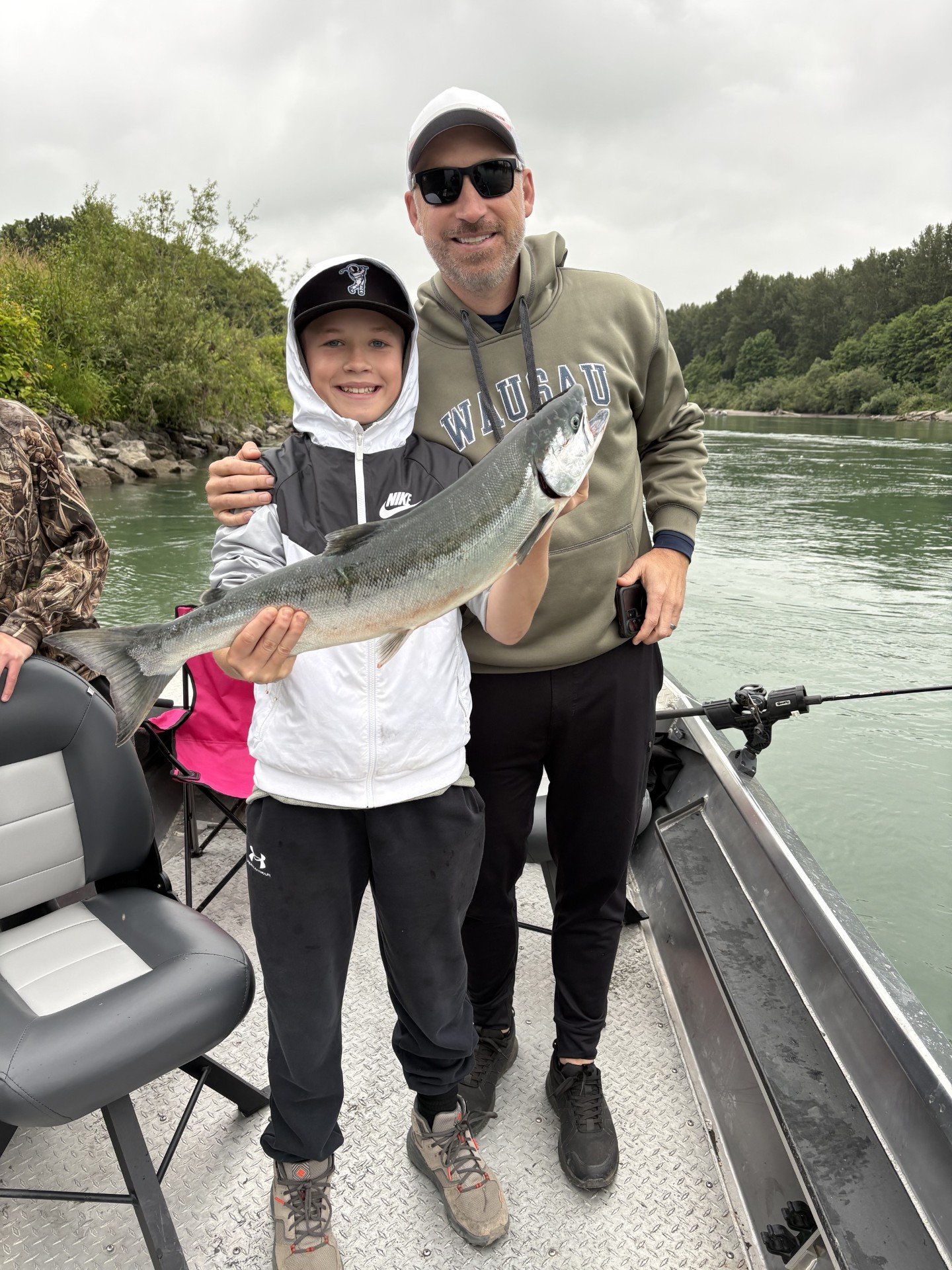 A smiling young boy and adult man on a boat, holding a large fish they caught, with water and trees in the background.