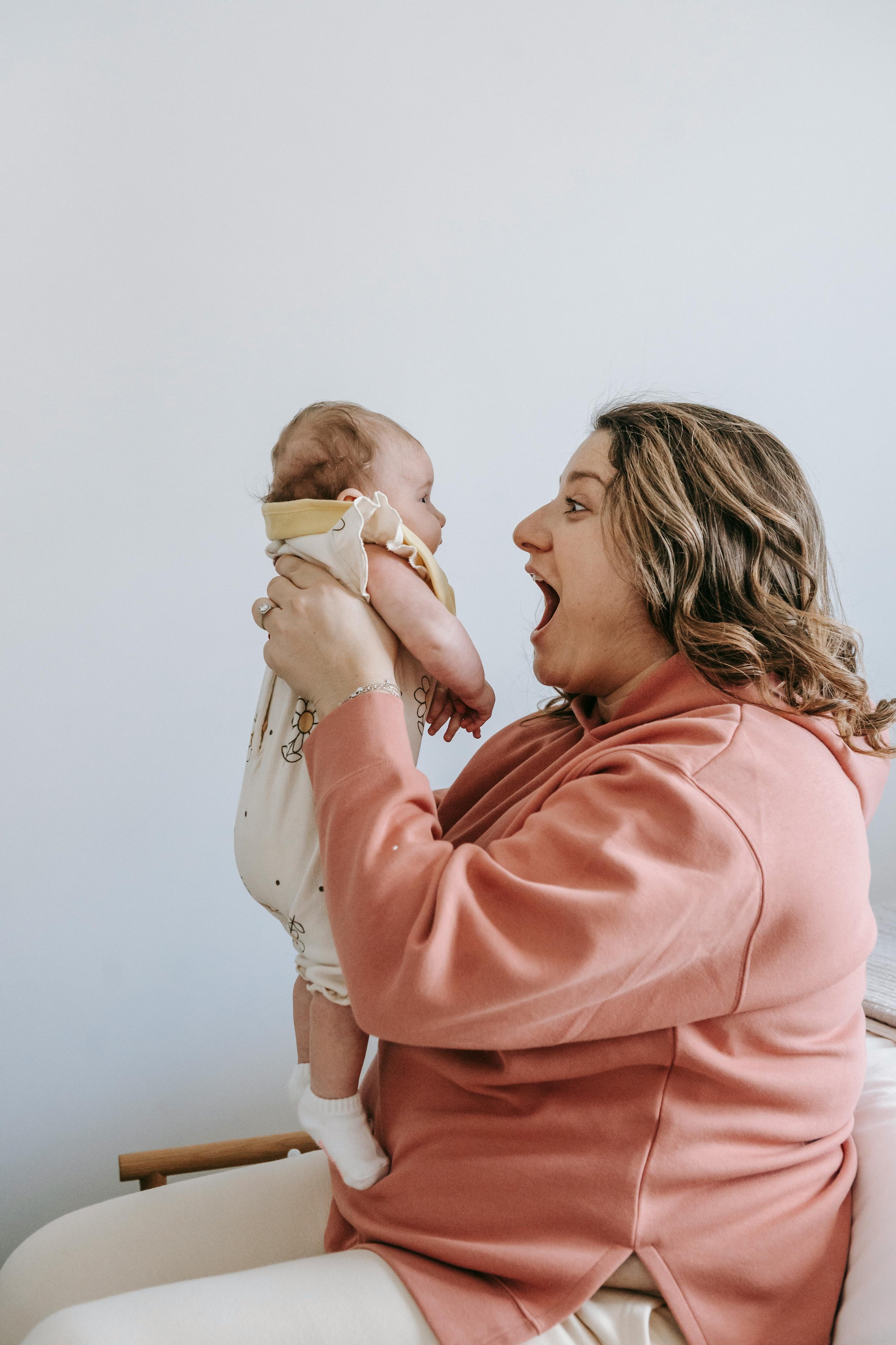 A woman with wavy brown hair wearing a pink hoodie holds a baby up in the air. The baby is dressed in a beige and yellow outfit with floral patterns and white socks, looking at the woman with an open mouth. The background is a plain white wall.
