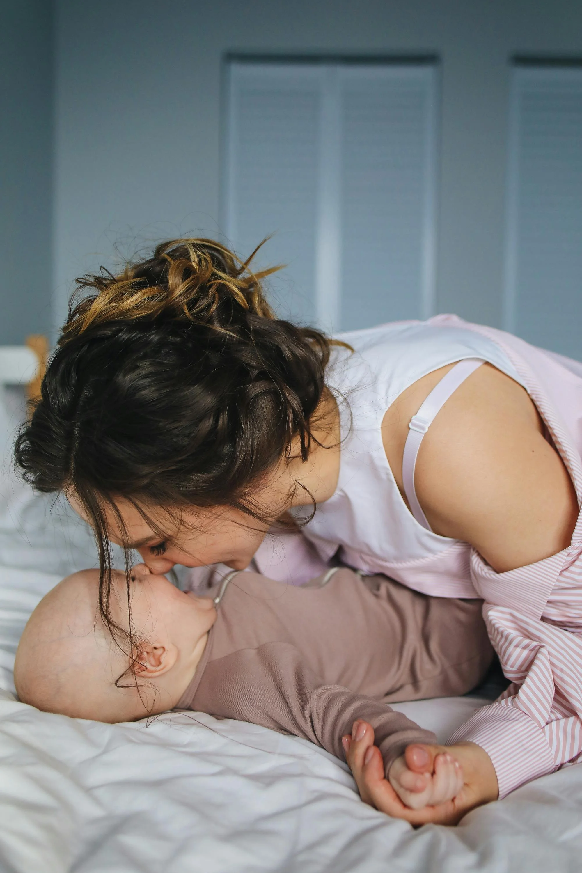 A woman with wavy hair leaning over a sleeping baby on a bed.